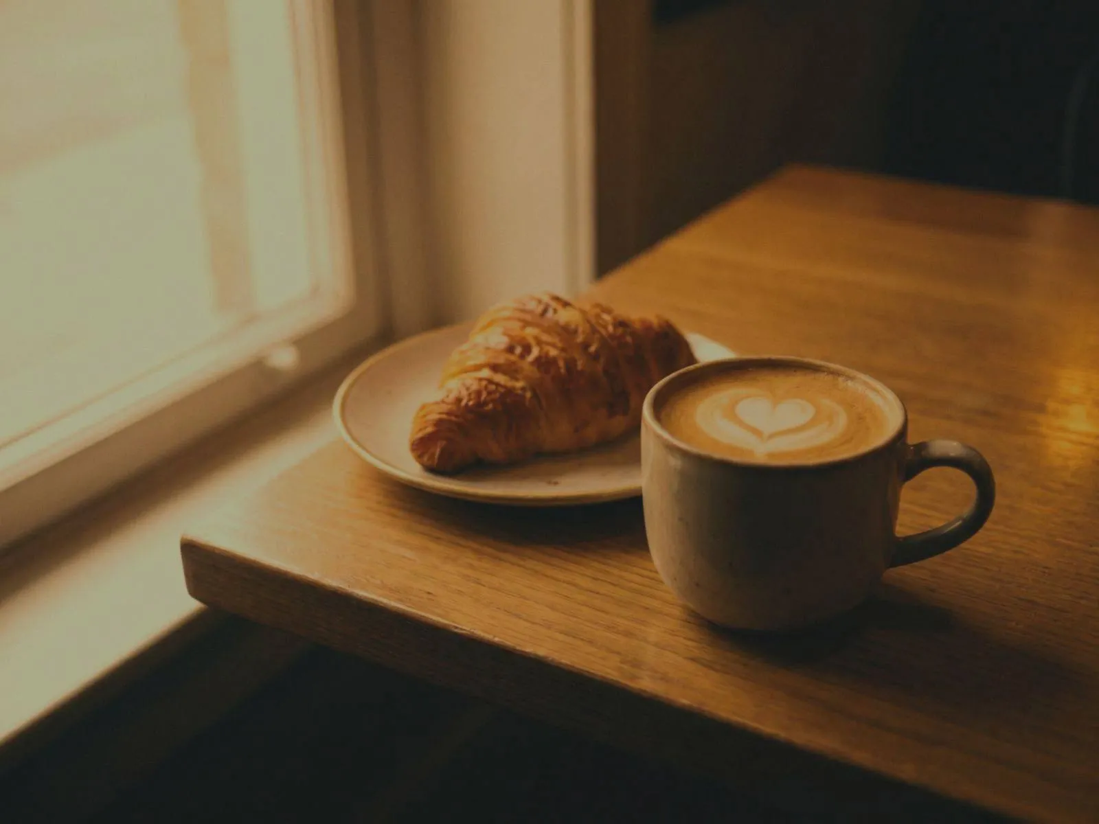A dim phone photo of a coffee and a croissant by a window